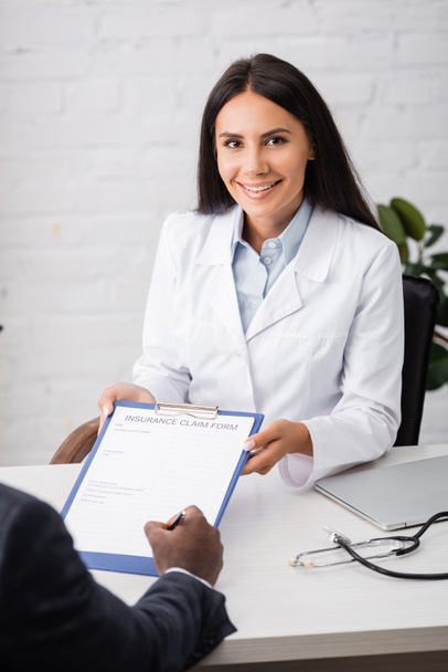 brunette, joyful doctor holding clipboard while african american patient signing insurance claim form  - Photo, image