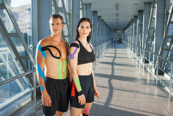 Portrait of muscular brunette woman with arms crossed and man looking at camera with strong face. Young couple athletes posing indoors, colorful kinesiotaping on body, futuristic interior. - Foto, imagen