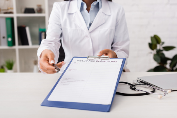 przycięty widok lekarza posiadającego formularz roszczenia ubezpieczeniowego w pobliżu stetoskopu na biurku - Zdjęcie, obraz