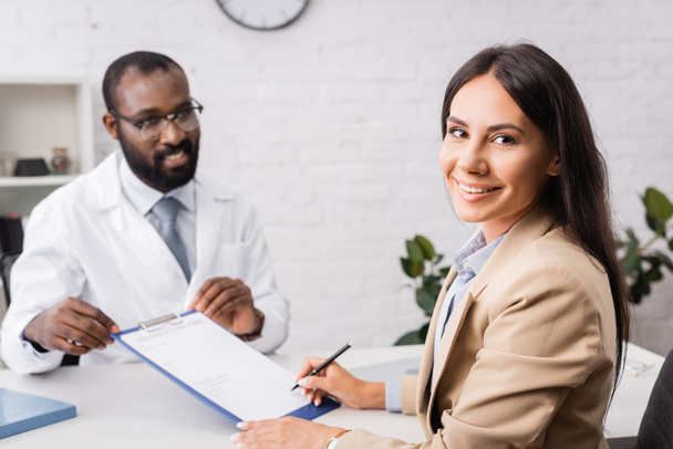 selektiver Fokus der freudigen Frau, die in die Kamera schaut, während sie in der Nähe eines amerikanischen Arztes ein Versicherungsformular unterschreibt - Foto, Bild