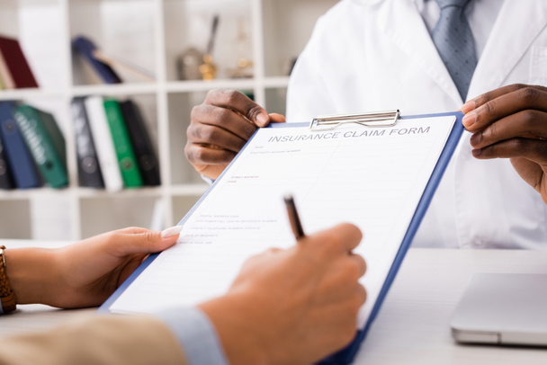 vue recadrée du patient signant le formulaire de demande de règlement d'assurance auprès du médecin afro-américain - Photo, image