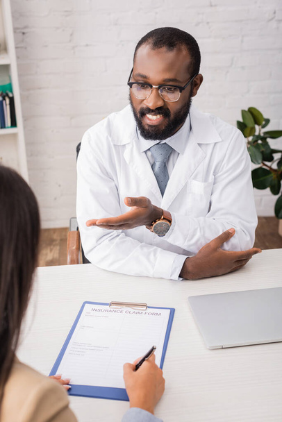 enfoque selectivo del médico afroamericano en gafas que apuntan con la mano cerca de firmar el formulario de reclamo de seguro del paciente - Foto, Imagen