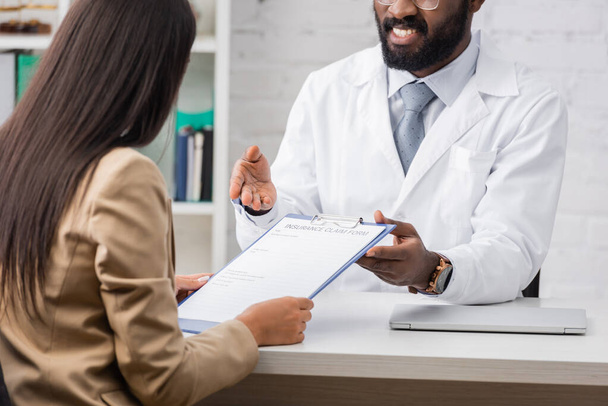 selektywne skupienie się afrykańskiego lekarza wskazując ręką na formularz roszczenia ubezpieczeniowego w pobliżu brunetka kobieta - Zdjęcie, obraz