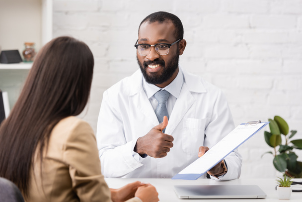 joyful african american doctor showing thumb up while holding clipboard with insurance form near brunette woman - Foto, Bild