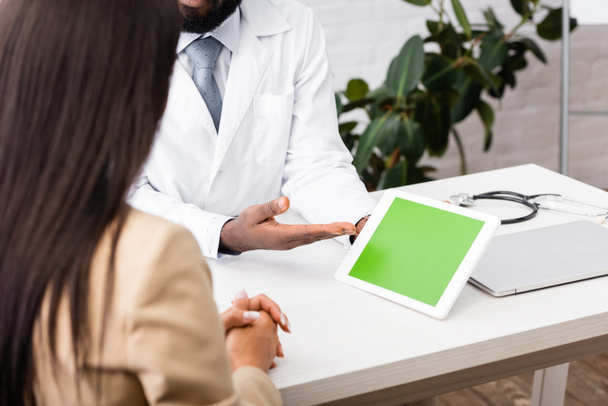 обрезанный вид африканского американского врача указывая рукой на цифровой планшет с зеленым экраном рядом с брюнеткой пациента - Фото, изображение