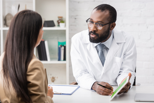 selective focus of serious african american doctor showing digital tablet while talking to brunette woman - Photo, Image