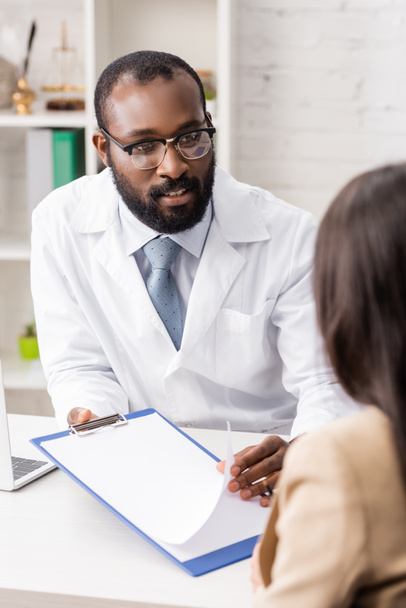 enfoque selectivo del médico afroamericano hablando con el paciente mientras sostiene el portapapeles con papel en blanco - Foto, imagen