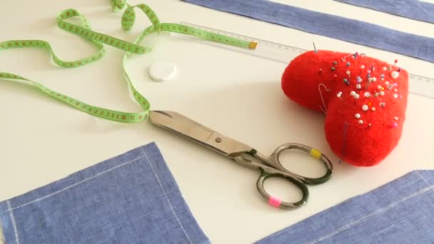 Kit da cucire, forbici da cucire, scarti di tessuto blu, un cuscino rosso con aghi a forma di cuore, un centimetro su uno sfondo bianco del tavolo. Laboratorio di cucito, flusso di lavoro della fabbrica del giocattolo - Filmati, video
