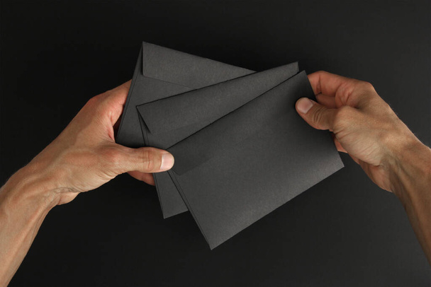 deux mains masculines caucasiennes tenant trois enveloppes noires vierges sur fond noir, maquette avec espace de copie  - Photo, image