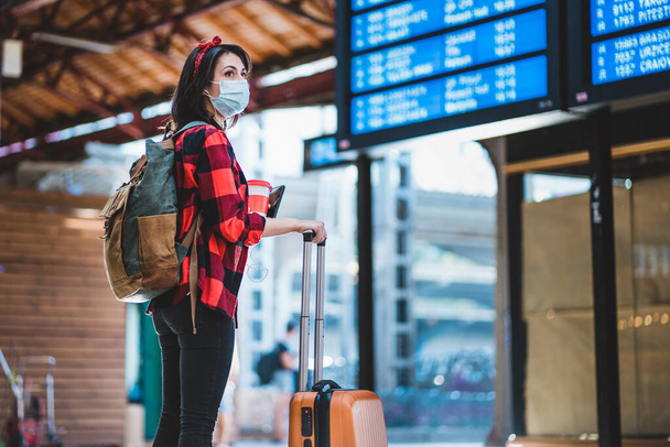 Mladá žena v ochranné lékařské masce stojí proti informační tabuli na mezinárodním letišti nebo vlakovém nádraží. Hipsterová samice s batohem a taškou na vozík hledá cestu. Pandemický koncept cestování - Fotografie, Obrázek