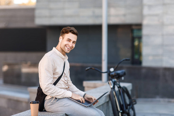 Networking outdoors. Happy guy in wireless headphones with coffee, works on laptop in city - Foto, imagen