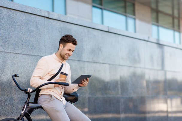 Modern lifestyle. Smiling guy with cup of coffee looks in tablet, sits on bike - Foto, Bild