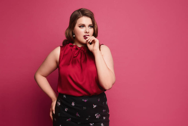 Pensive plus-size model girl with bright makeup and with stylish hairstyle in a red blouse and skirt posing at the pink background, isolated - Photo, Image