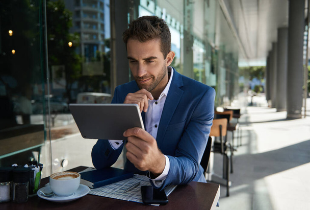 Joven empresario sonriente bebiendo café y usando una tableta digital mientras está sentado afuera en una mesa de café - Foto, Imagen