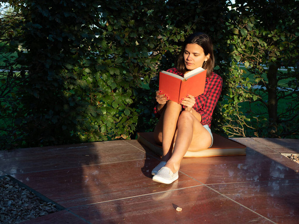 Giovane ragazza adulta caucasica in camicia rossa si siede nel parco al mattino e legge libro - Foto, immagini