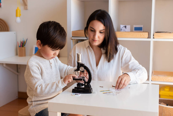 Lillte Kid explorando a ciência com um microscópio e ajuda da mãe ou do professor. A passear em casa. Comunidade de aprendizagem. Montessori Schoo - Foto, Imagem