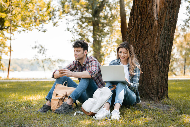 couple using laptop and smartphone while sitting under tree trunk  - Fotó, kép