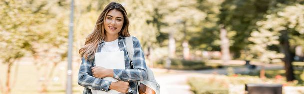 panoramic shot of woman in plaid shirt holding laptop and looking at camera in park  - Фото, изображение