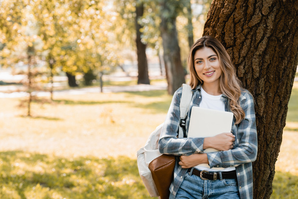 woman in plaid shirt holding laptop and looking at camera near tree trunk  - Foto, Bild