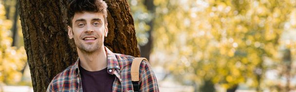 panoramic orientation of man in checkered shirt looking at camera near tree trunk  - Photo, image
