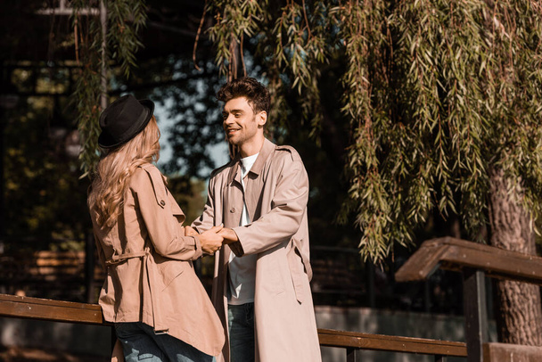 man in trench coat looking at woman in hat while holding hands in park - Photo, Image