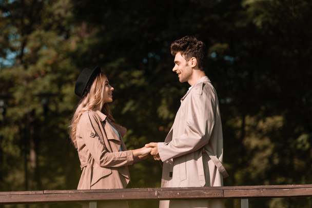 side view of stylish couple in trench coats holding hands in park - Photo, Image