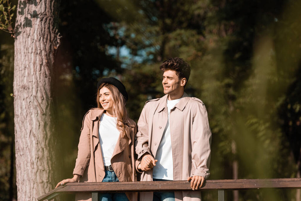 stylish couple in trench coats looking away and holding hands in park - Photo, Image