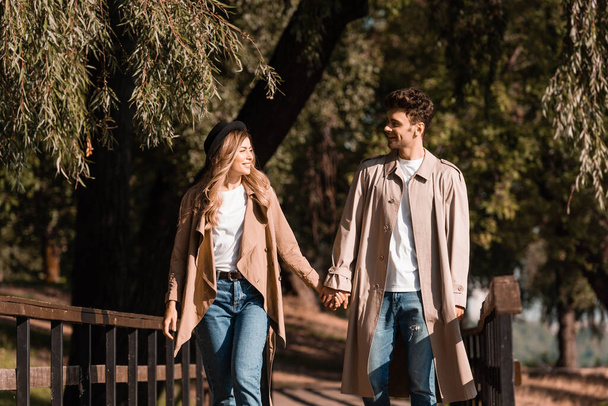 couple in trench coats holding hands, looking at each other and walking on wooden bridge - Photo, Image