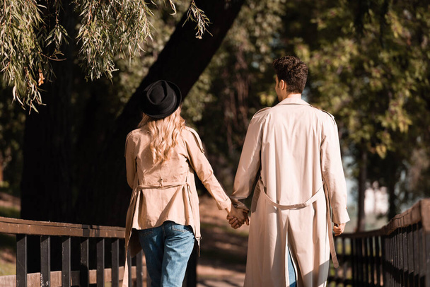 back view of couple in trench coats holding hands and walking on wooden bridge in autumnal park - Foto, immagini