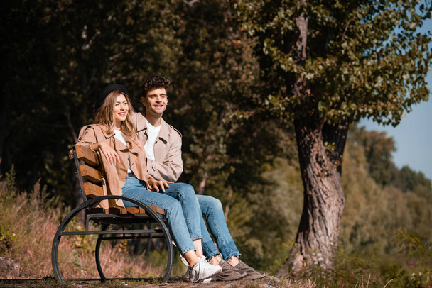 blonde woman in hat and man in trench coat sitting on bench in park - Foto, Bild
