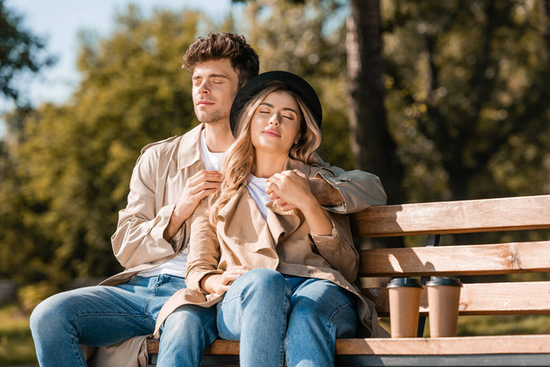 blonde woman in hat and man in trench coat sitting on bench near paper cups  - Photo, Image