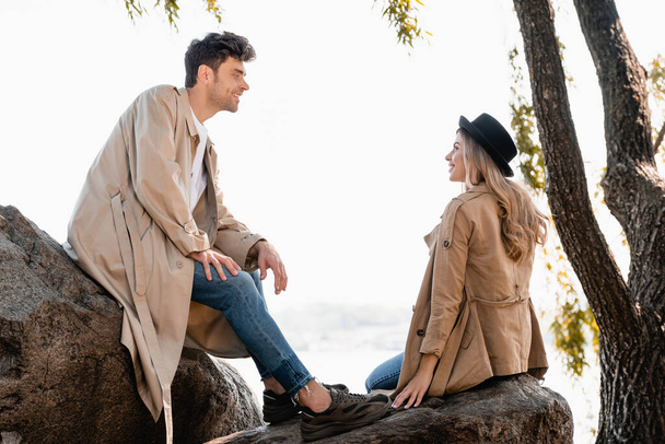 blonde woman in hat and man in trench coat sitting on stones and looking at each other - Foto, Imagen