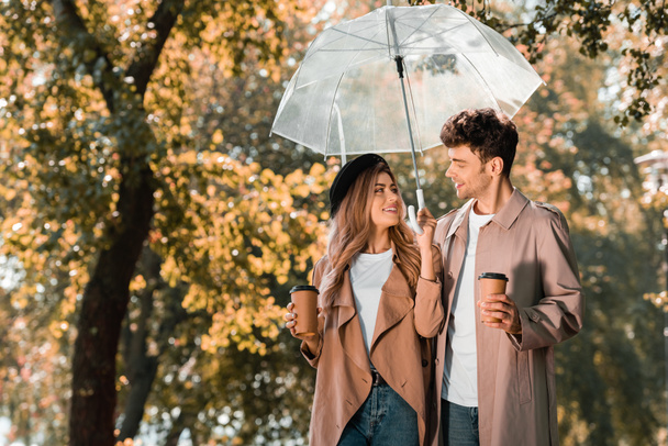 ζευγάρι σε καμπαρντίνες στέκεται κάτω από ομπρέλα και κρατώντας χάρτινα φλιτζάνια με καφέ για να πάει - Φωτογραφία, εικόνα