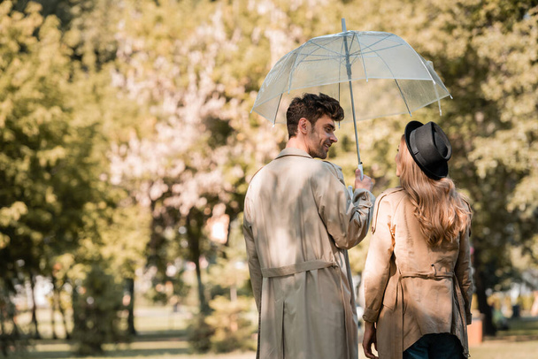 mujer en sombrero y hombre en gabardina de pie bajo el paraguas mientras se miran en el parque otoñal  - Foto, Imagen