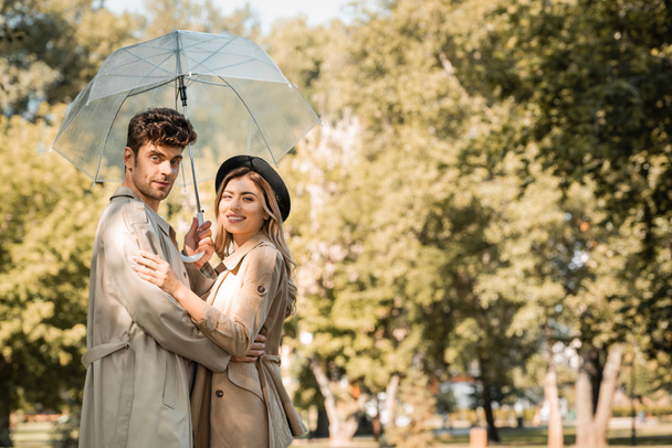 blonde woman in hat and man in trench coat hugging under umbrella and looking at camera in autumnal park  - Photo, Image
