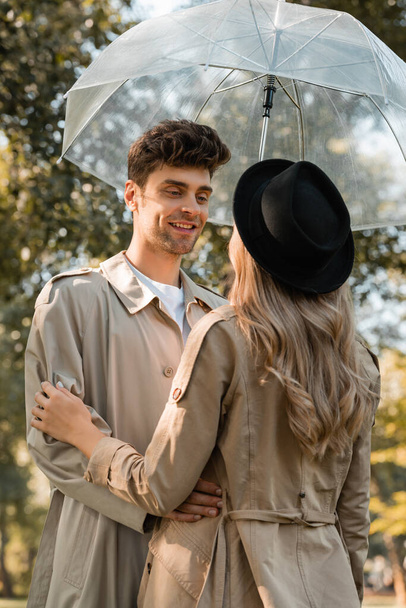 man in trench coat standing under umbrella and looking at woman in black hat in autumnal park  - Photo, Image