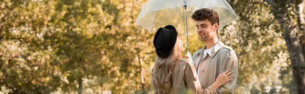 panoramic crop of woman in hat and man in trench coat standing under umbrella in autumnal park  - Photo, Image