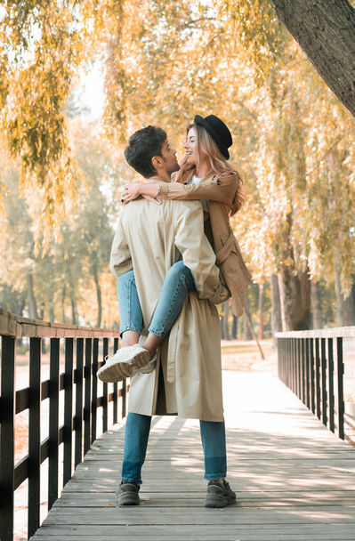 man in trench coat holding in arms woman in hat while standing on wooden bridge in autumnal park  - Φωτογραφία, εικόνα