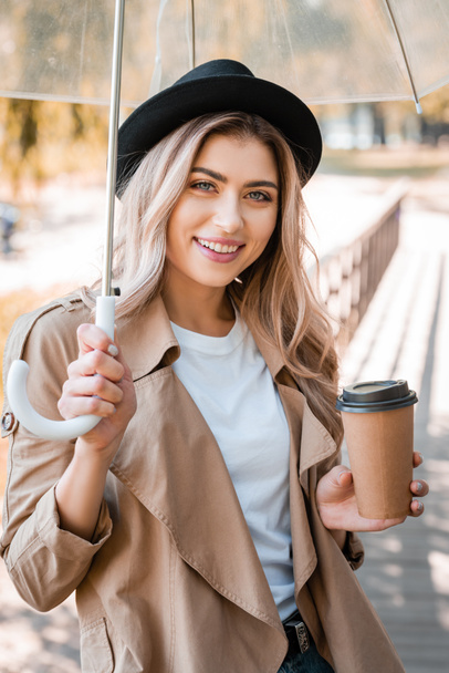 woman in hat holding umbrella and paper cup with coffee to go in autumnal park  - Фото, зображення