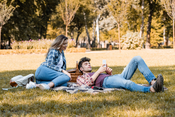 Frau und Mann schauen auf Smartphone, während sie sich auf Decke im Park ausruhen - Foto, Bild