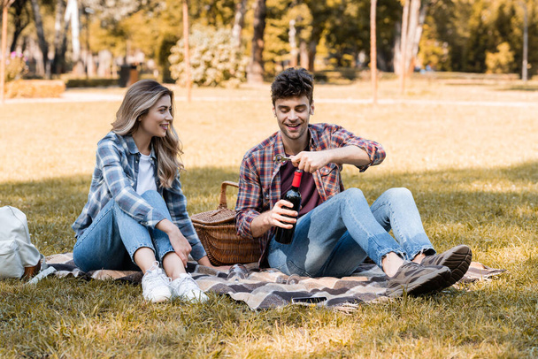 Kadının yanında elinde kırmızı şarap şişesi olan bir adam parkta battaniyede oturuyor.  - Fotoğraf, Görsel