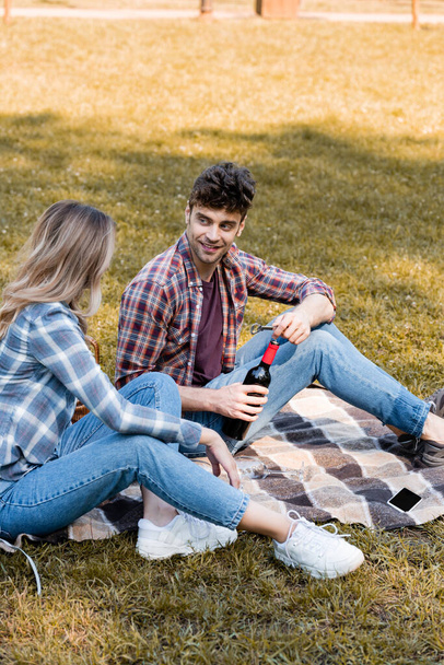 uomo in possesso di bottiglia di vino rosso vicino alla donna seduto sulla coperta plaid vicino smartphone nel parco  - Foto, immagini
