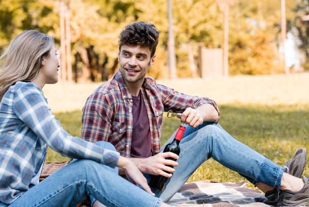 чоловік тримає пляшку червоного вина біля жінки, сидячи на плетеній ковдрі в парку
  - Фото, зображення