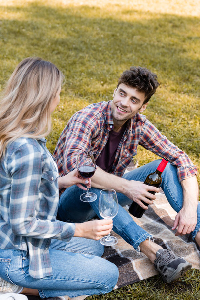 man holding bottle of red wine and looking at woman with glasses sitting on plaid blanket in park  - Foto, Imagem