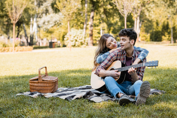woman sitting on plaid blanket near wicker basket and touching boyfriend playing acoustic guitar  - Photo, Image