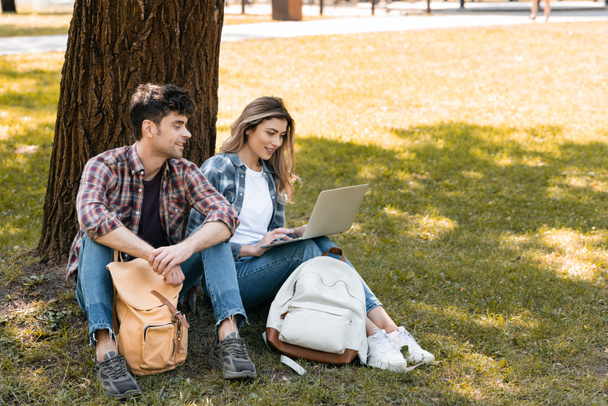 man op zoek naar laptop terwijl zitten in de buurt vrouw onder boomstam - Foto, afbeelding