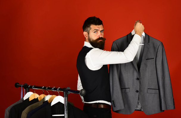 Tailor with serious face holds grey suit near custom jackets - Φωτογραφία, εικόνα
