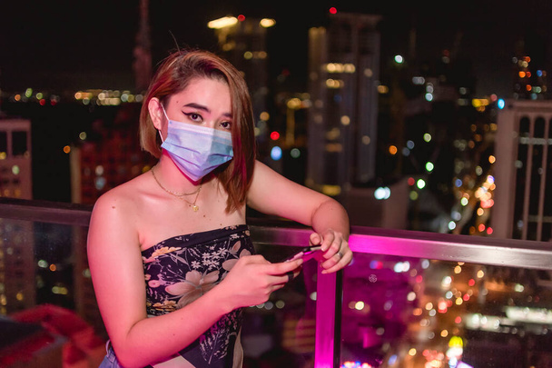 Atrakcyjna młoda Azjatka w masce chirurgicznej patrzy w kamerę trzymając telefon w barze na dachu w Makati, Metro Manila. - Zdjęcie, obraz