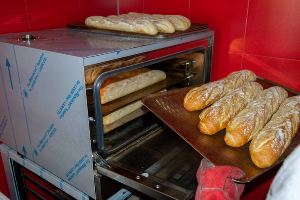 Pekař odstraní bochníky chleba z trouby, jakmile jsou již upečené a s opékanou barvou s červenými rukavicemi - Fotografie, Obrázek