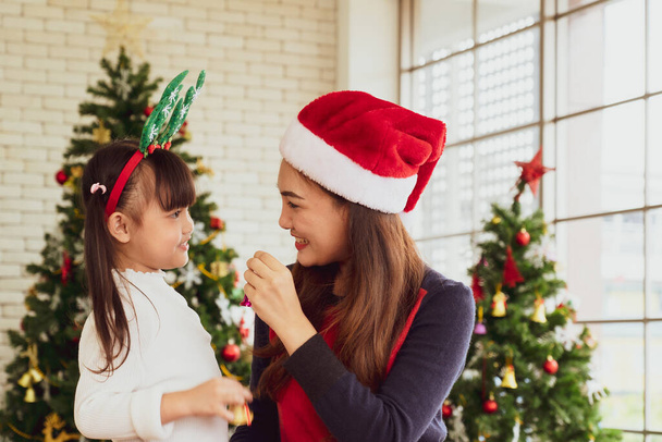 アジア系の母親と若い娘はサンタの帽子をかぶってクリスマスツリーを飾るために飾りを使用しています。家族みんなでコンセプト - 写真・画像